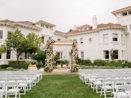 A sunny wedding setup in a garden courtyard with white chairs, floral arches, and a white mansion backdrop, ready for vows.