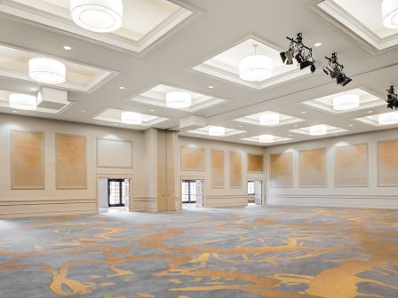 A bright empty banquet hall with recessed lights, patterned carpet, and stage rigging hanging from the ceiling.