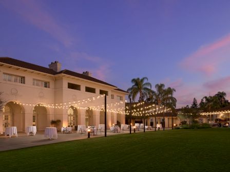 A courtyard event setup at dusk with an arched building, string lights, tables with white cloths, chairs, and palm trees.