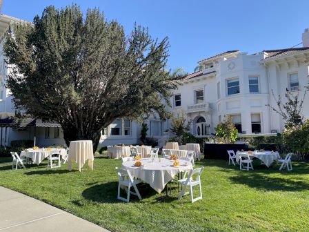 Outdoor dining setup with white tablecloths on a green lawn in front of a white villa under a clear blue sky.