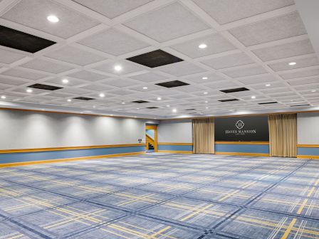 An empty event hall with patterned carpet, bright recessed lights, and a stage backdrop reading &ldquo;Hayes Mansion, San Jose.&rdquo;