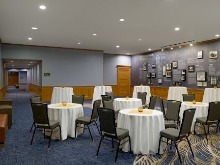 A banquet-style meeting room with round tables, chairs, and a history exhibit wall of framed photos.