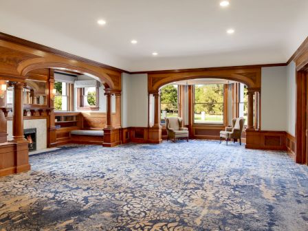 An elegant living room with ornate wooden archways, a fireplace, patterned blue carpet, two armchairs, and large windows with greenery.