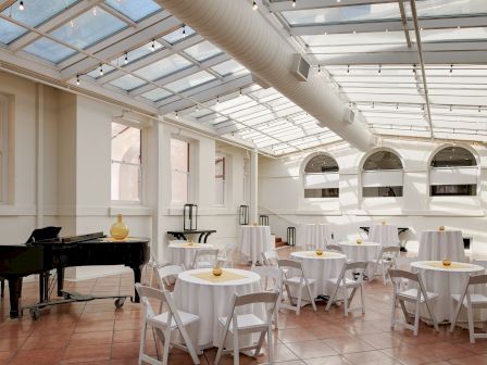A bright modern event hall with arched windows, string lights, round tables with white cloths and candles, and a grand piano.