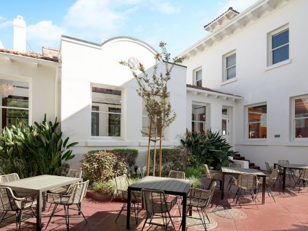 A bright courtyard patio with white stucco houses, arched entry, garden plants, and outdoor tables with chairs under sunny skies.