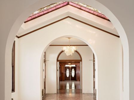 A bright hallway inside a building with arched doorways, a marbled floor, and hanging chandeliers under a stained-glass ceiling.