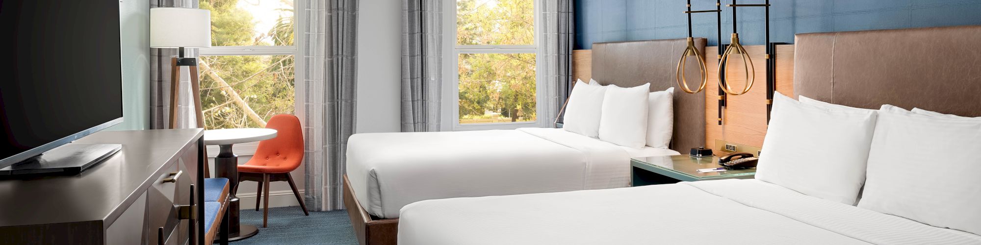 A modern hotel room with two white beds, blue accent wall, and large windows with gray curtains, overlooking greenery.