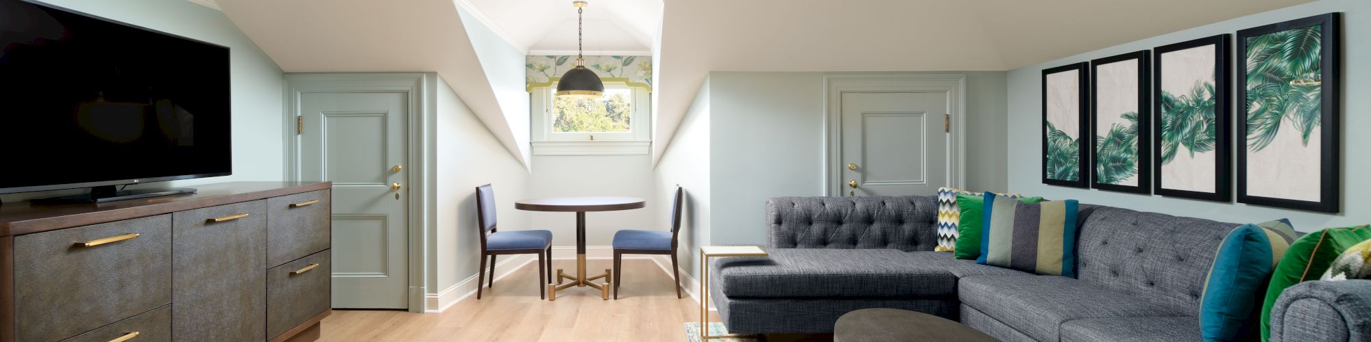 Bright living room with gray sectional, TV, colorful pillows, framed tropical art, and a small round table under a skylight.