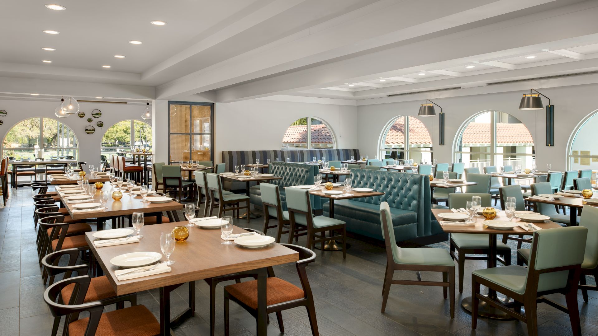 Bright restaurant dining room with neatly set tables, cushioned chairs, and large arched windows looking out to trees.