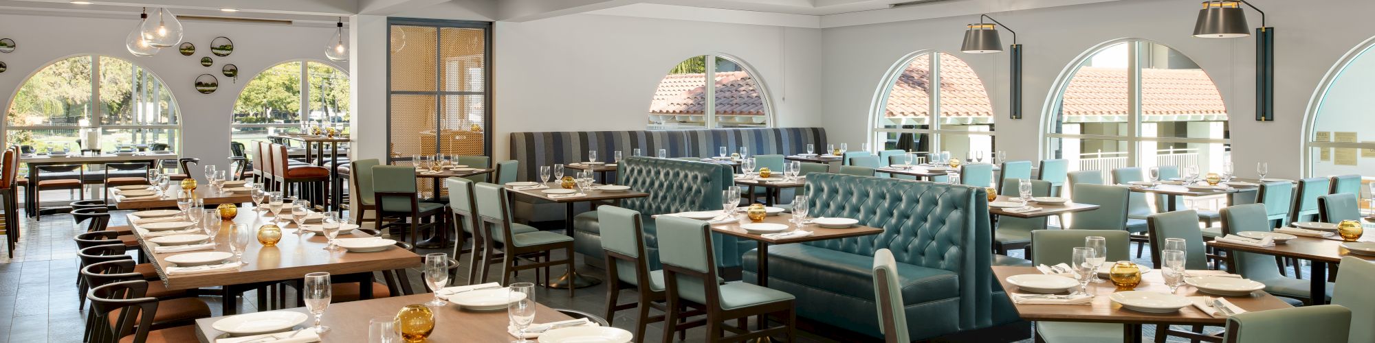 Bright restaurant dining room with neatly set tables, cushioned chairs, and large arched windows looking out to trees.