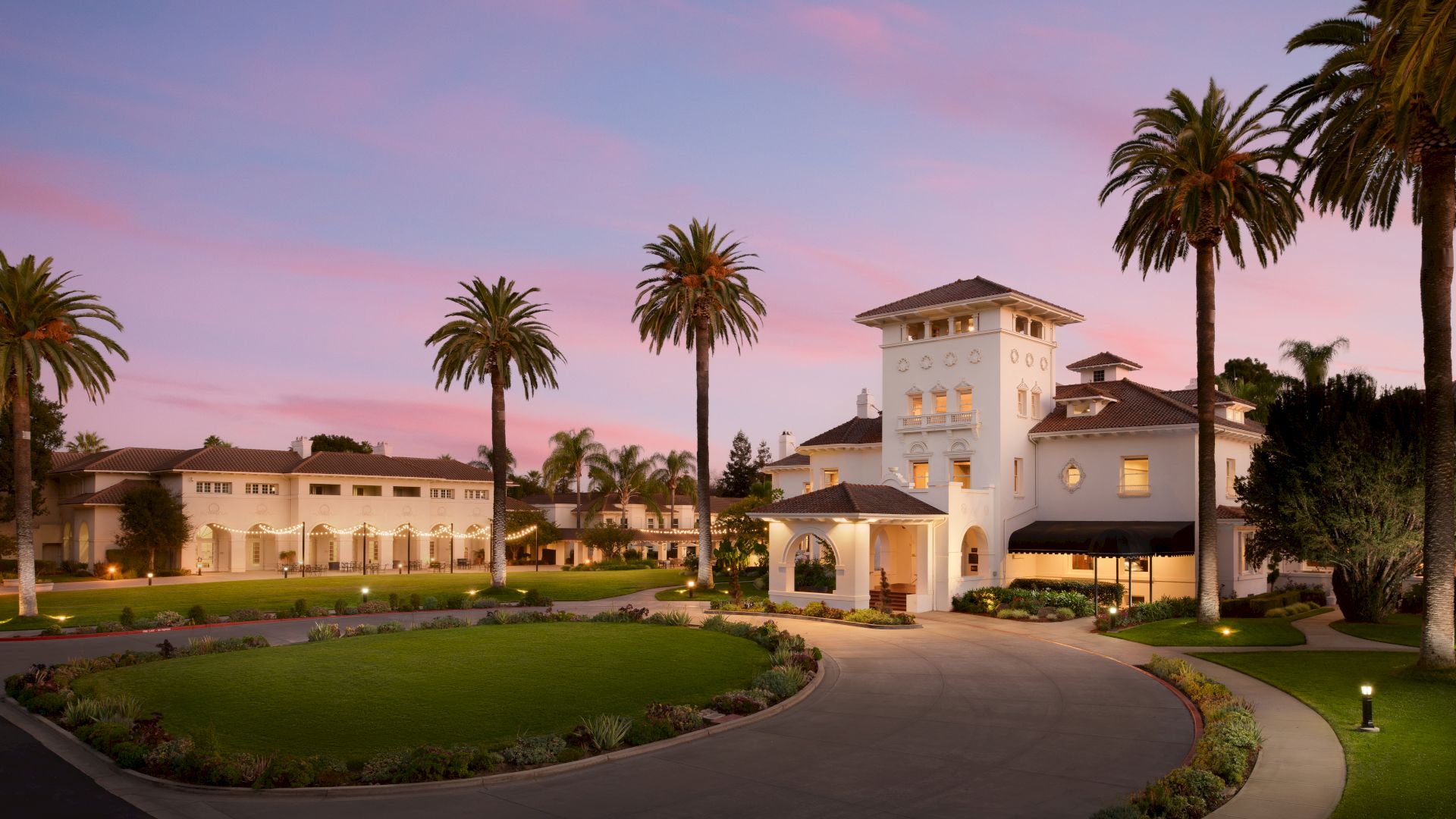 A white villa/hotel courtyard at dusk, lit pathways and arches, surrounded by tall palm trees against a pink-purple sky.