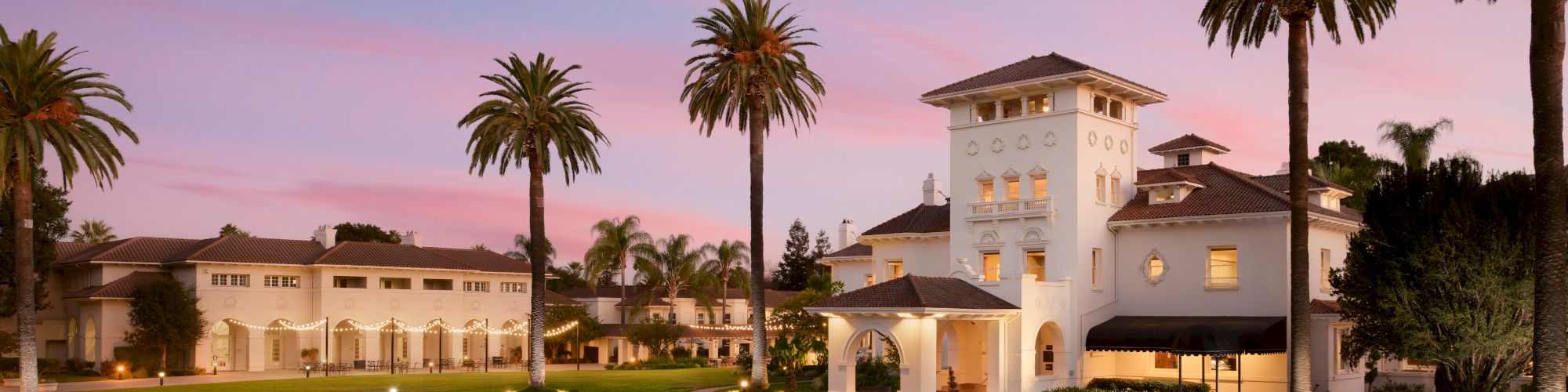 A white resort mansion with arched balconies and warm lights, surrounded by tall palm trees at sunset under a pink-purple sky.