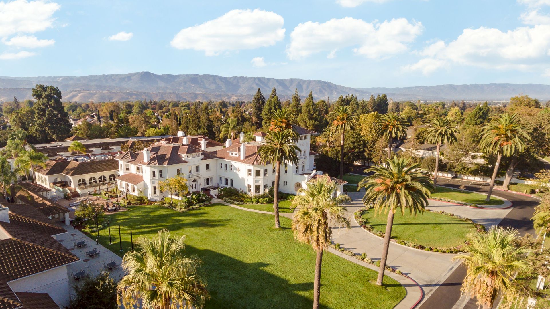 Aerial view of a large white villa with tiled roofs, palm-lined gardens, winding paths, and distant mountains under blue sky.
