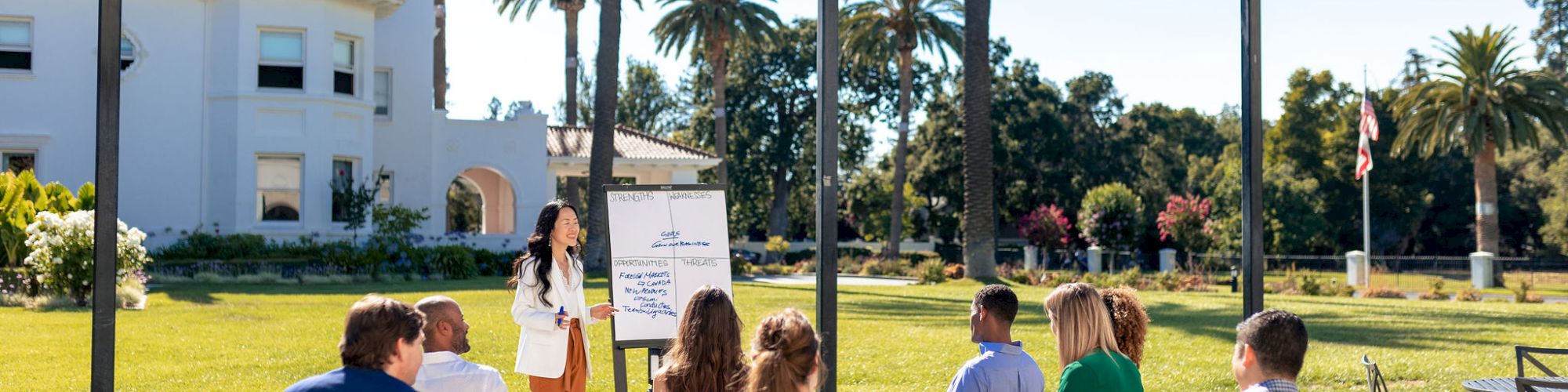 An outdoor seminar: a presenter talks to people seated on chairs by a whiteboard under string lights and palm trees.