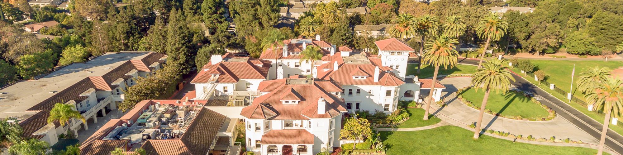 Aerial view of a luxury estate with red-tile homes, manicured gardens, palm trees, and a blue pool overlooking hills and suburbs.
