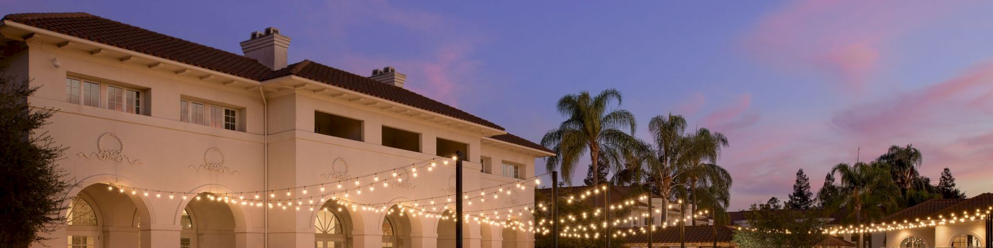 An outdoor courtyard event space with white chairs and tables under warm string lights as the sun sets and palm trees sway.
