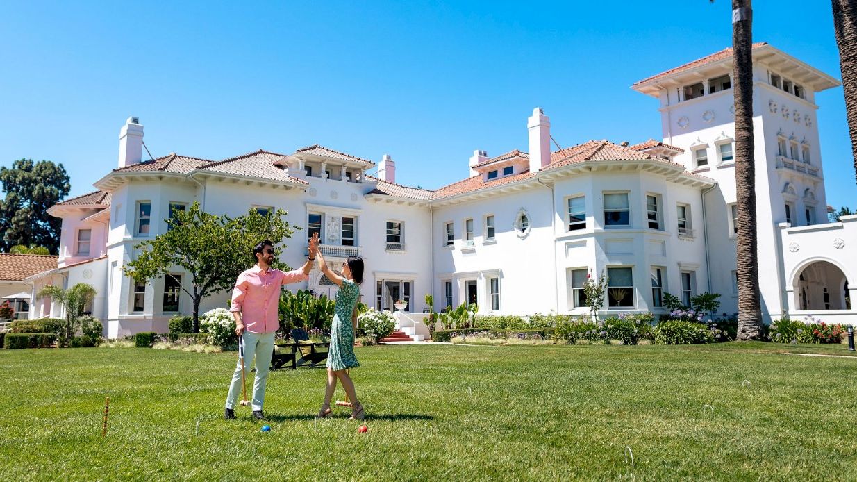 A man and woman play on a sunny lawn in front of a large white Mediterranean-style mansion with palm trees.