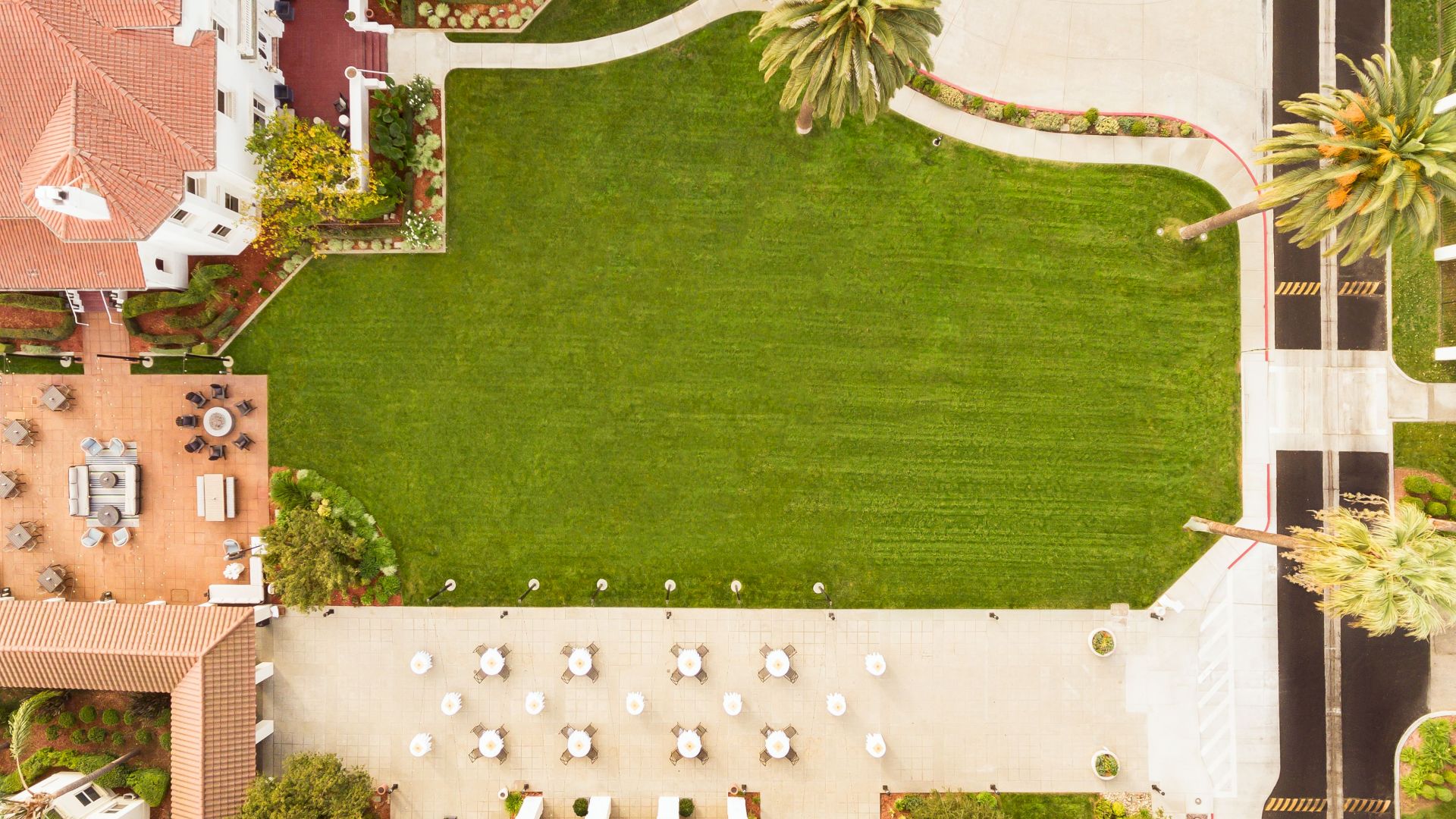 Aerial view of a landscaped resort courtyard with a large green lawn, curving paths, palm trees, and terrace seating.