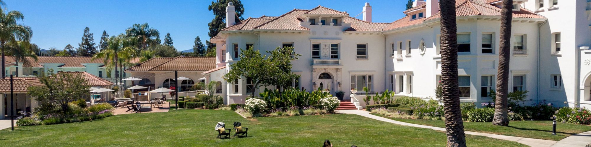 A sunny courtyard with a green lawn, palm trees, and a large white stucco building, with two people walking nearby.