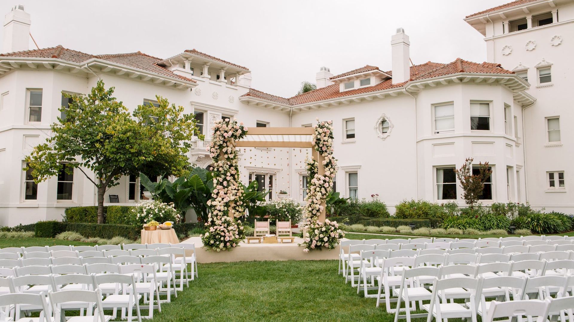 Outdoor wedding ceremony setup with white chairs, floral arch, and decorated stage in front of a white mansion.