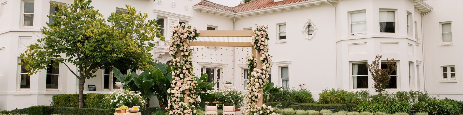 Outdoor wedding ceremony setup with white chairs, floral arch, and decorated stage in front of a white mansion.