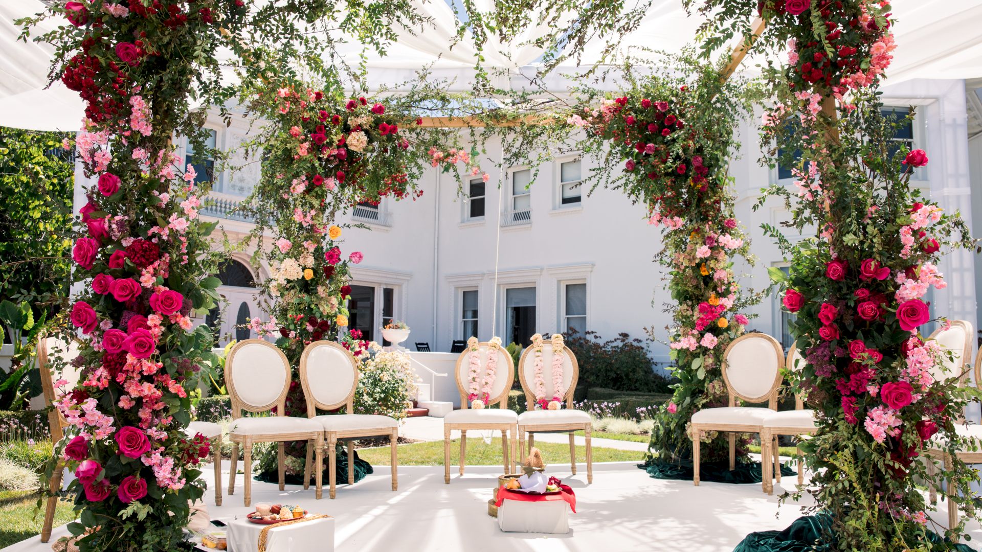 Floral wedding backdrop under a white tent: hanging roses, a rose arch canopy, and decorated chairs on the garden aisle.