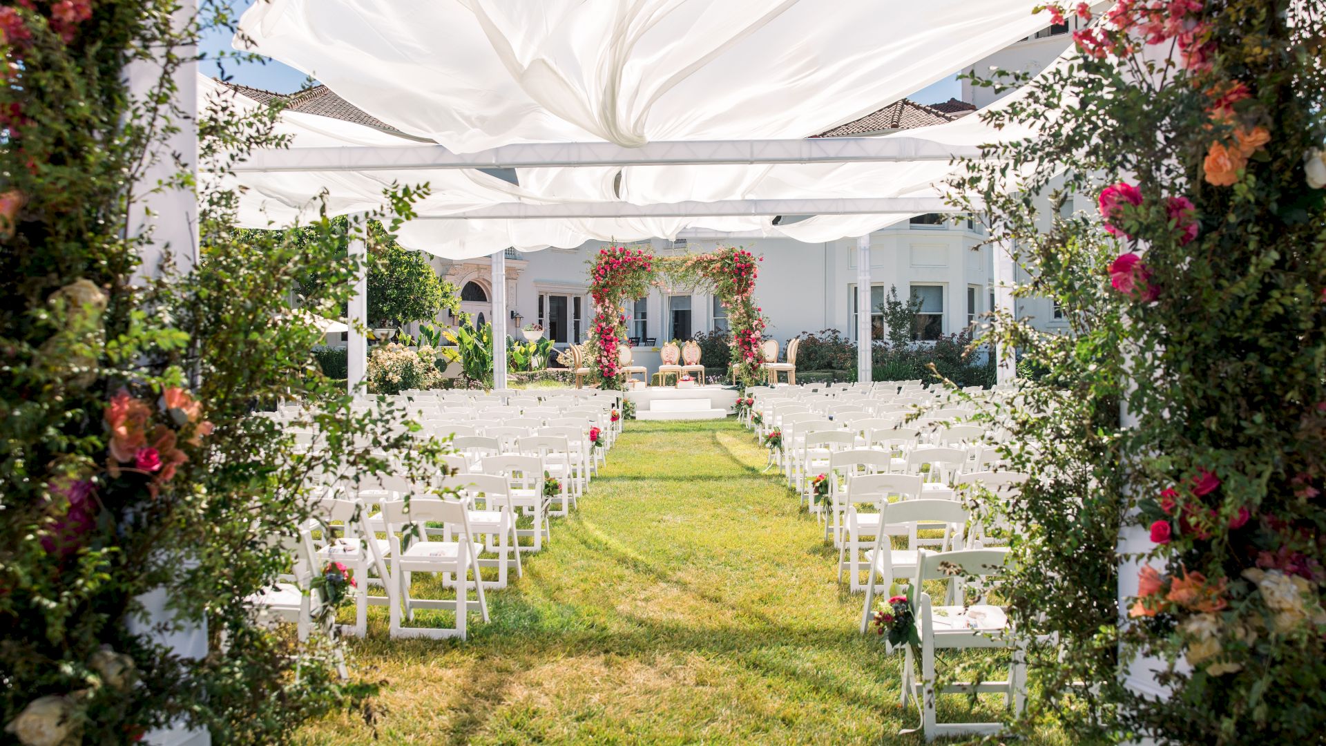 A garden wedding ceremony setup with a white canopy, rows of chairs, and pink flower arches around a sunny lawn.