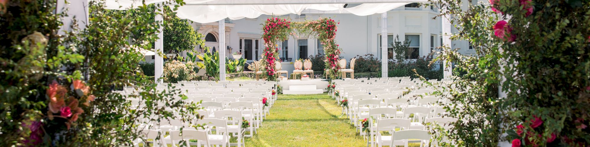 A garden wedding ceremony setup with a white canopy, rows of chairs, and pink flower arches around a sunny lawn.