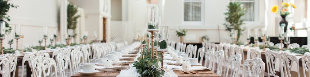 A bright wedding hall with white chairs, long tables, greenery centerpieces, and flowers in a sunlit conservatory.