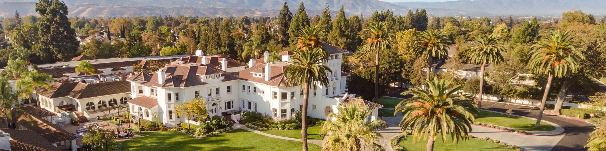 Aerial view of a large white resort building with manicured lawns, palm trees, winding paths, and distant mountains under a sunny sky.