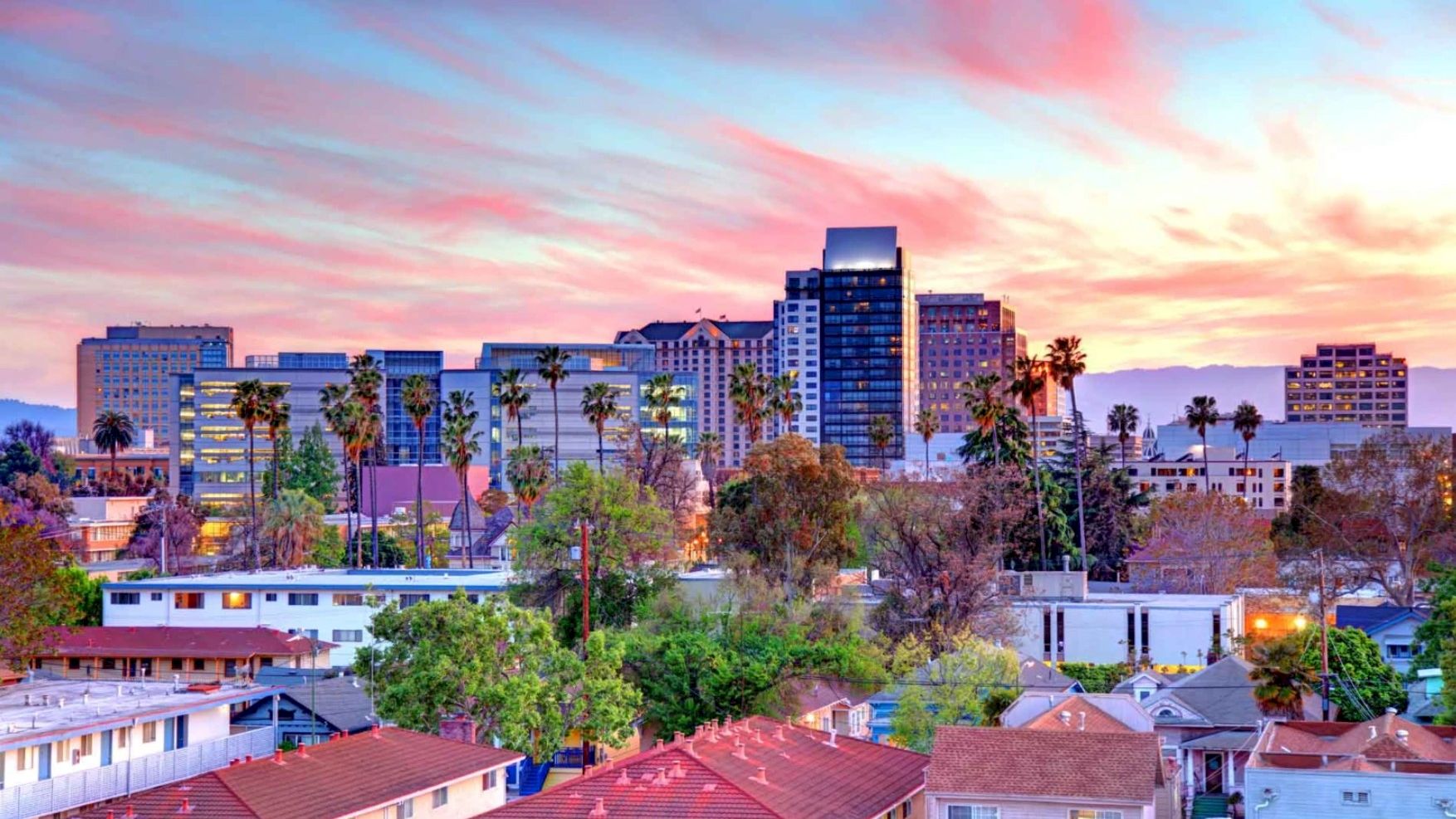 City skyline with palm trees and mid-rise buildings at sunset, glowing pink-orange clouds behind.