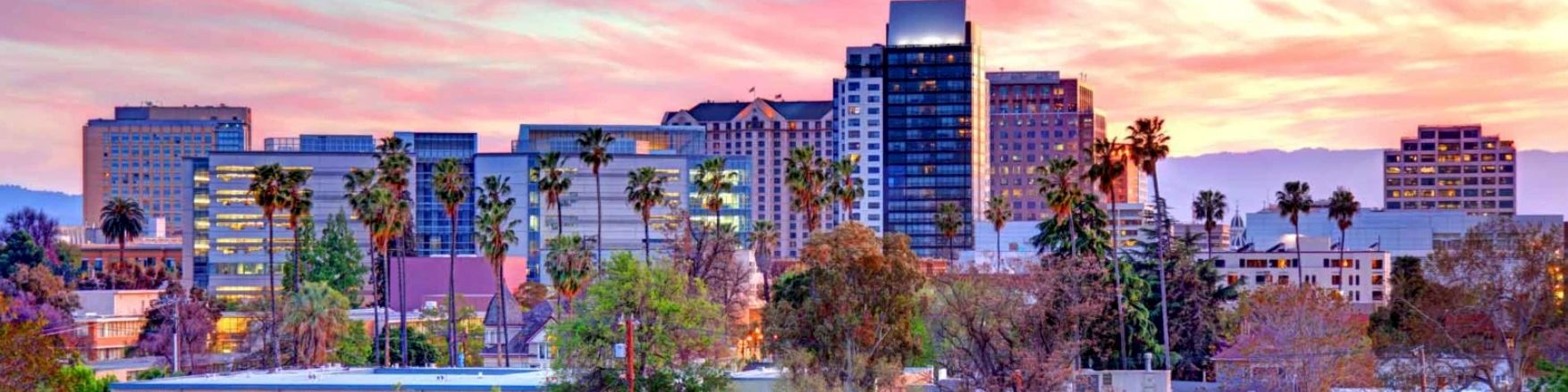 City skyline with palm trees and mid-rise buildings at sunset, glowing pink-orange clouds behind.