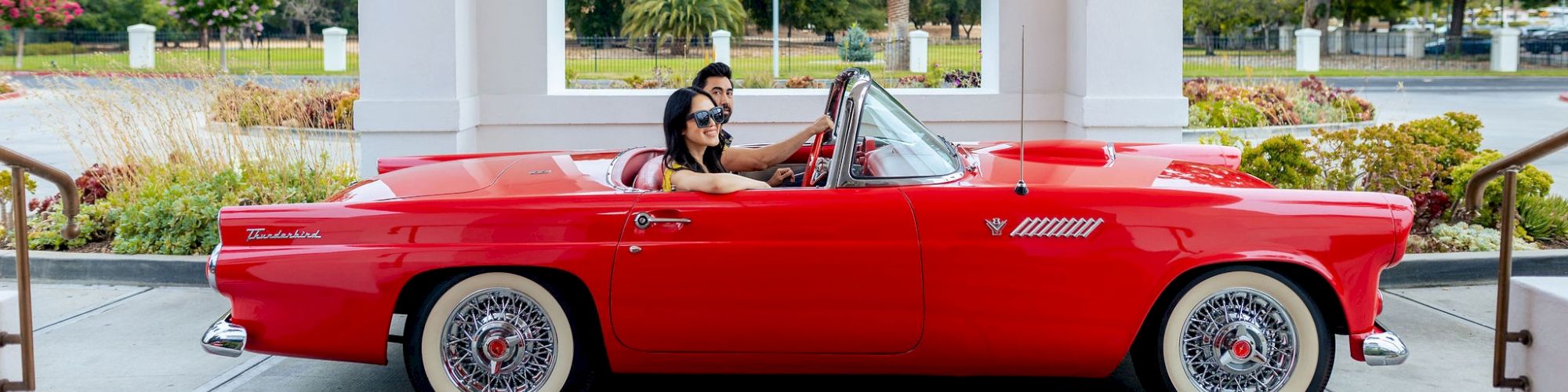 A bright red vintage Thunderbird convertible parked by landscaped gardens, with a driver and passenger posing under palm trees.