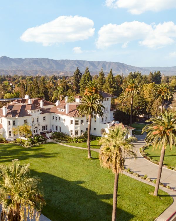 A sunny campus or resort with well-kept lawns, palm trees, white buildings, and a winding driveway under a blue sky with fluffy clouds.