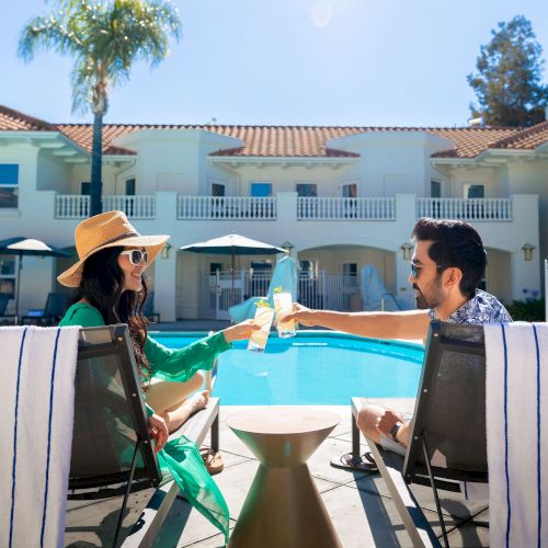 Two people toast by a pool at a sunny resort, lounging on striped chairs with a palm tree and a white building in the background.
