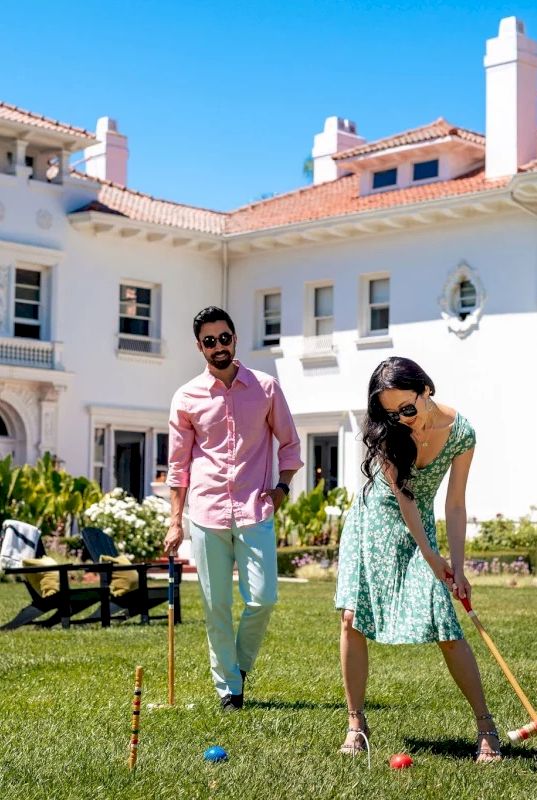 Two people play a lawn game outdoors in a sunny courtyard of a white building, aiming wooden mallets and colorful balls.