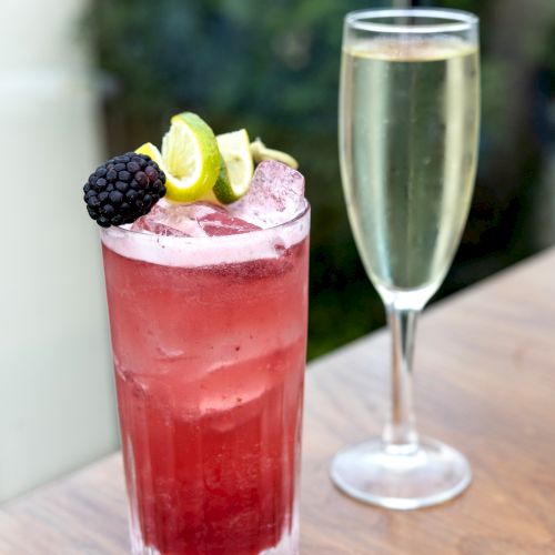 A tall red berry cocktail with ice and blackberry-lime garnish sits on a table beside an empty champagne flute.