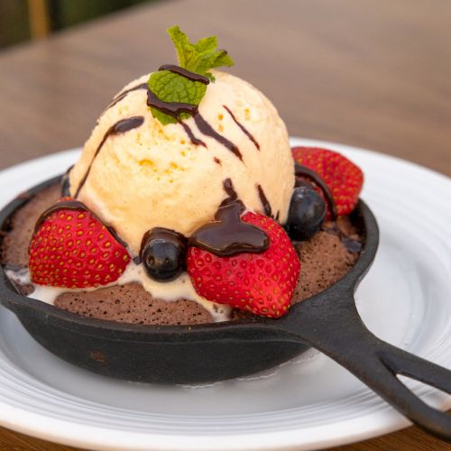 A bowl of vanilla ice cream with chocolate drizzle, strawberries, blueberries, and a mint leaf garnish.