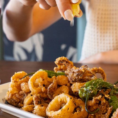 A plate of fried calamari and onion rings, with a lemon wedge being squeezed, a glass of red wine nearby, and a person in the background.
