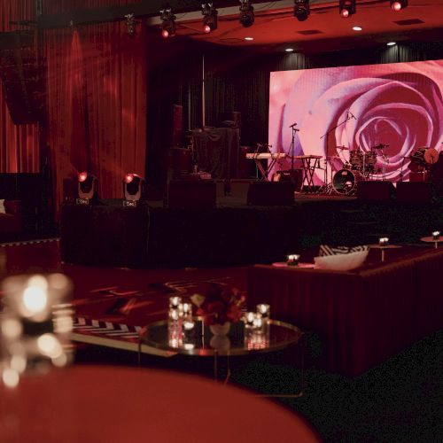 A dimly lit upscale lounge with red velvet seating, candlelit tables, and a stage featuring instruments; a swirling pink rose backdrop fills a large screen.