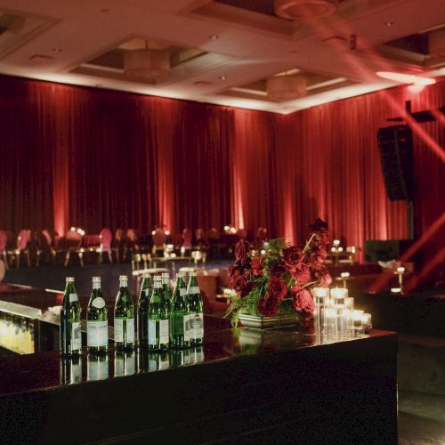 A sleek bar with bottled waters, candles, and floral arrangements set against a dim, red-lit stage backdrop.