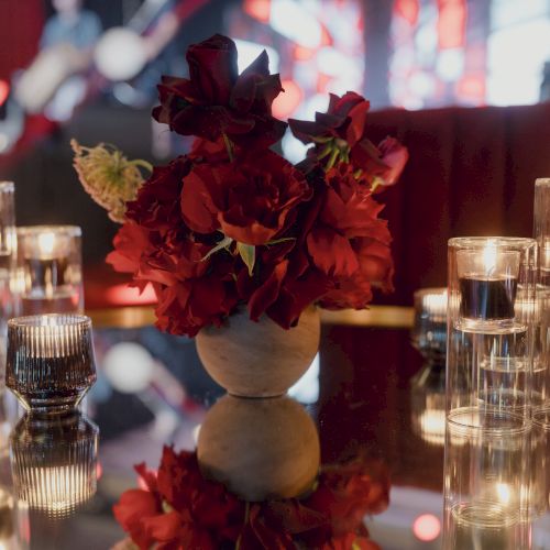 A centerpiece of red flowers in a white vase on a glass table, surrounded by lit candles in tall glass holders, creating a warm, romantic glow.