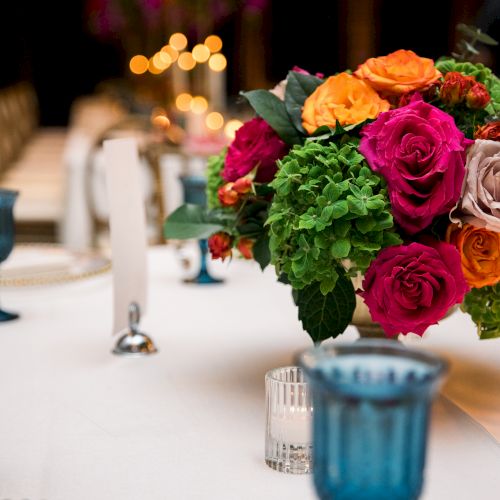 A table centerpiece with a vibrant bouquet of roses in orange, pink, and purple, surrounded by blue glassware and candles.