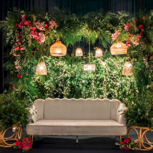 A vintage beige sofa framed by a lush green backdrop with hanging wicker lamps, florals, and candles for a cozy, garden-style backdrop.