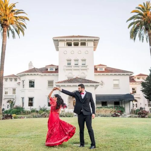 A couple dances joyfully on a manicured lawn in front of a grand white mansion with tall palm trees on either side, under a clear sky.