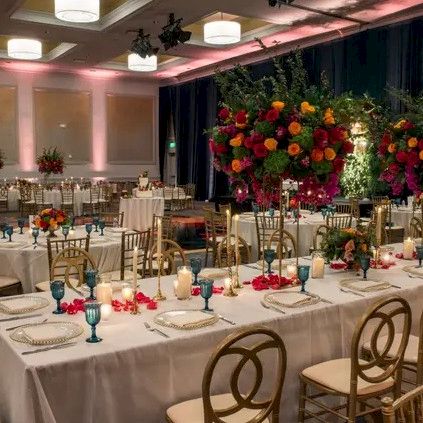 A formal banquet setup with long tables, white linens, elegant place settings, and large colorful flower arrangements in a dimly lit event hall. Top it at 140 characters, always ending the sentence.