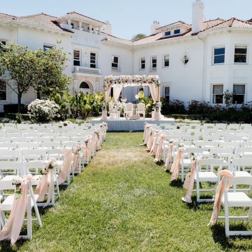 A sunny outdoor wedding setup with rows of white chairs and a floral arch ceremony on a lawn in front of a white building.