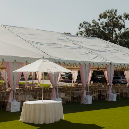 An outdoor event setup: a large white tent with pink drapes, chairs, and decorated tables on a lawn.