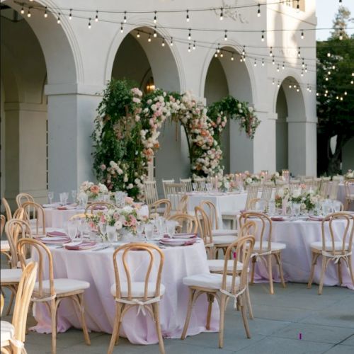 Outdoor wedding reception with round tables, pastel linens, wooden chairs, string lights, and floral arches under arches.