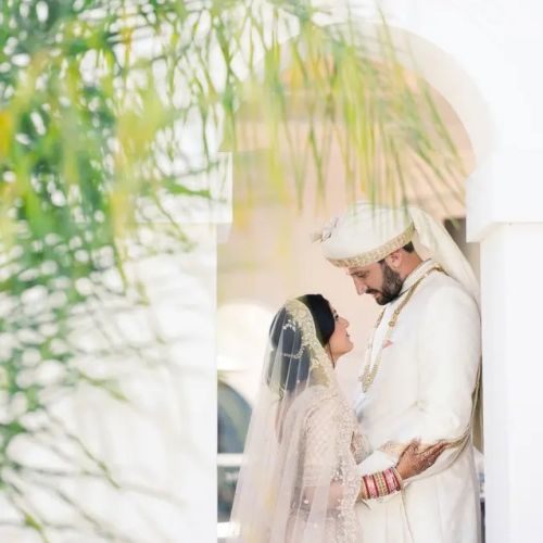 Two people in wedding attire (bride and groom) sharing a kiss/close moment in a bright window niche, with greenery framing the scene.